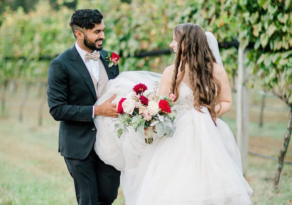 Bride and groom walk in vineyard, bridal bouquet of red and white roses with dusty gray hints in greenery.
