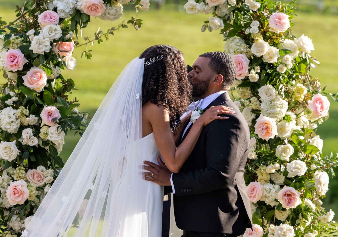 black bride and groom kissing in front of flower arch made of pale pink and white roses, summer wedding at Veritas